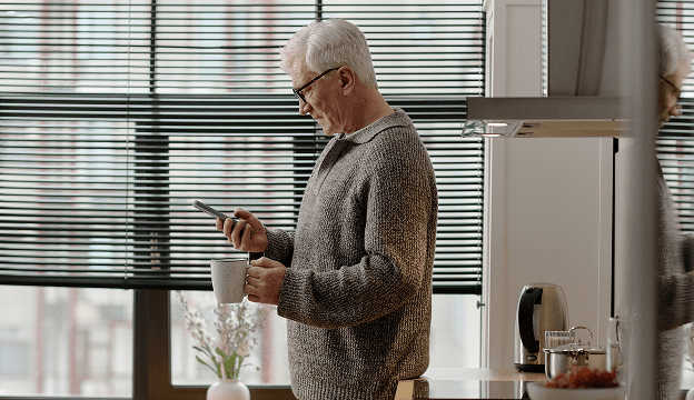 An older man standing in his kitchen holding a smart phone and a cup of coffee.