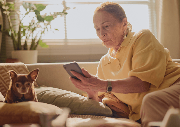 An older woman sitting on a couch looking at her phone, with a small dog beside her.