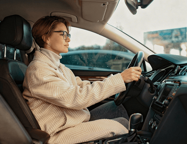 A female driver in a beige coat confidently behind the wheel of a vehicle.